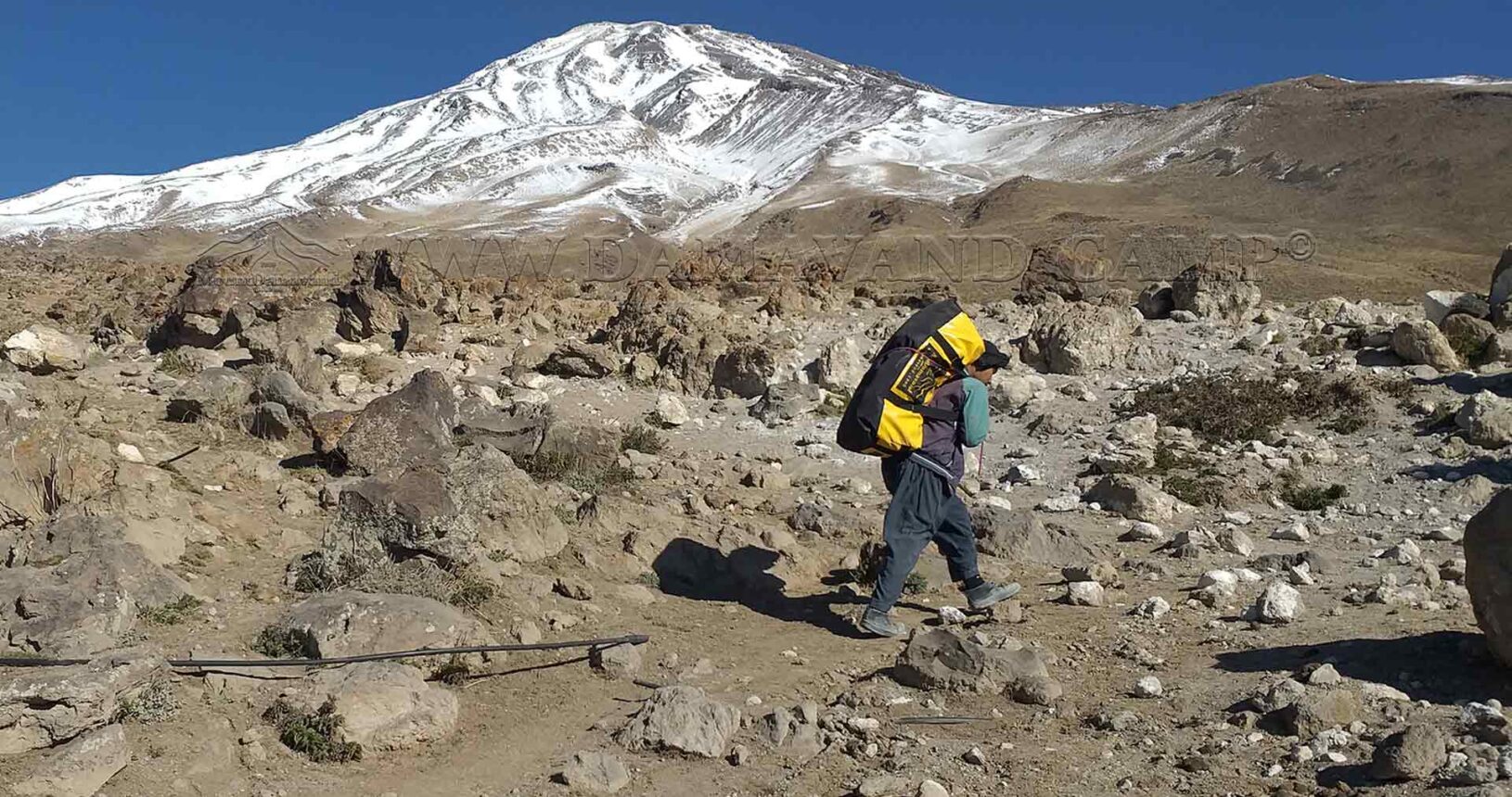 A dedicated porter carries a duffel bag, navigating the rugged terrain at the foothills of the majestic Mount Damavand.